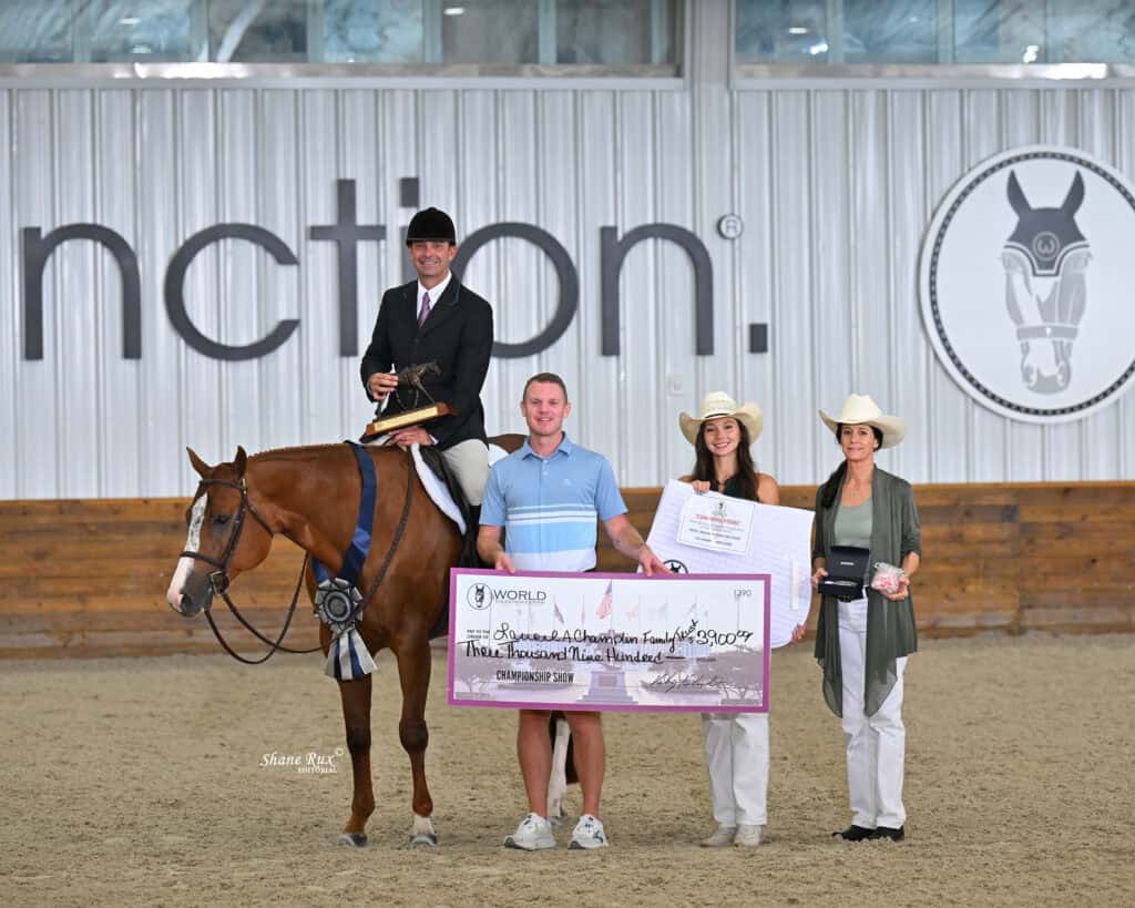 Four people and a horse stand indoors; one person is mounted. Two people hold a large check, and one holds a certificate. An arena wall with signage is visible in the background.