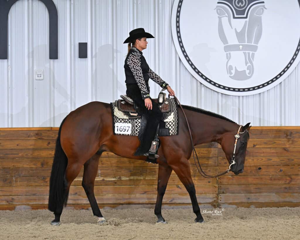 A person in western attire rides a brown horse inside an arena with wood paneling and a large horse logo on the wall.