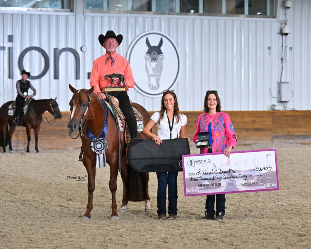 A man on horseback holds a trophy next to two women, one with a guitar case and one with an oversized check, inside an indoor riding arena.