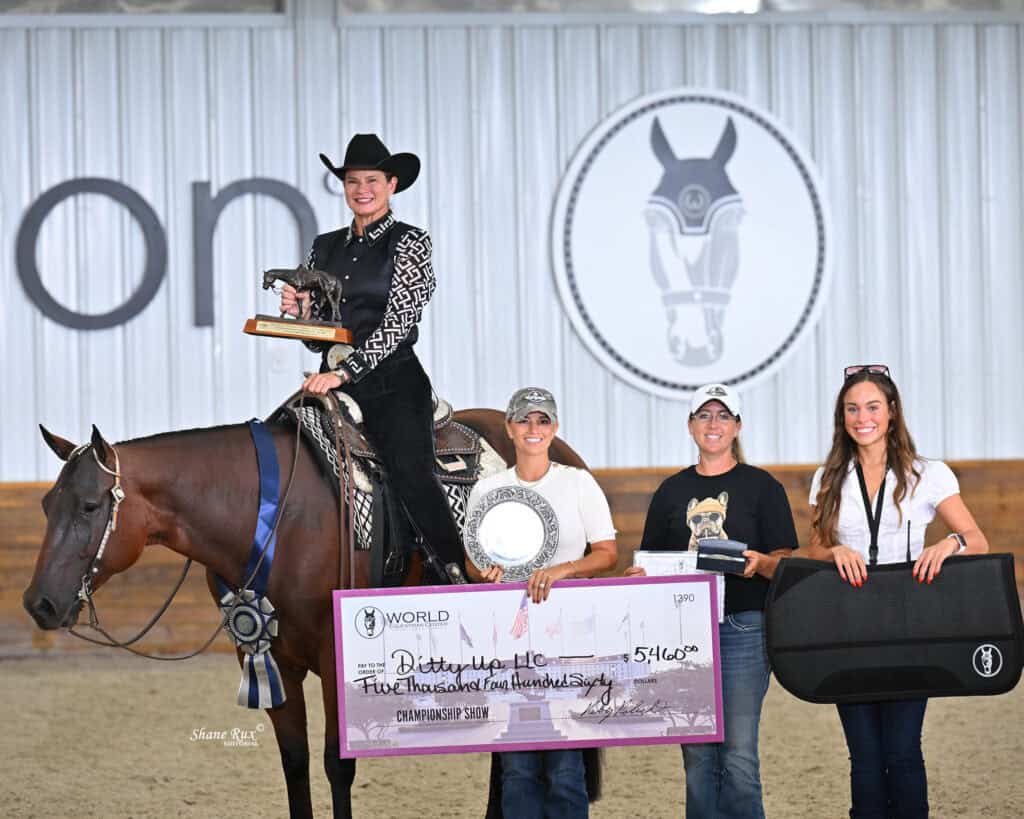 Four women pose with a horse, a trophy, and a large $5,460 check at an indoor equestrian event. One woman sits on the horse holding the trophy, while three stand in front, smiling.