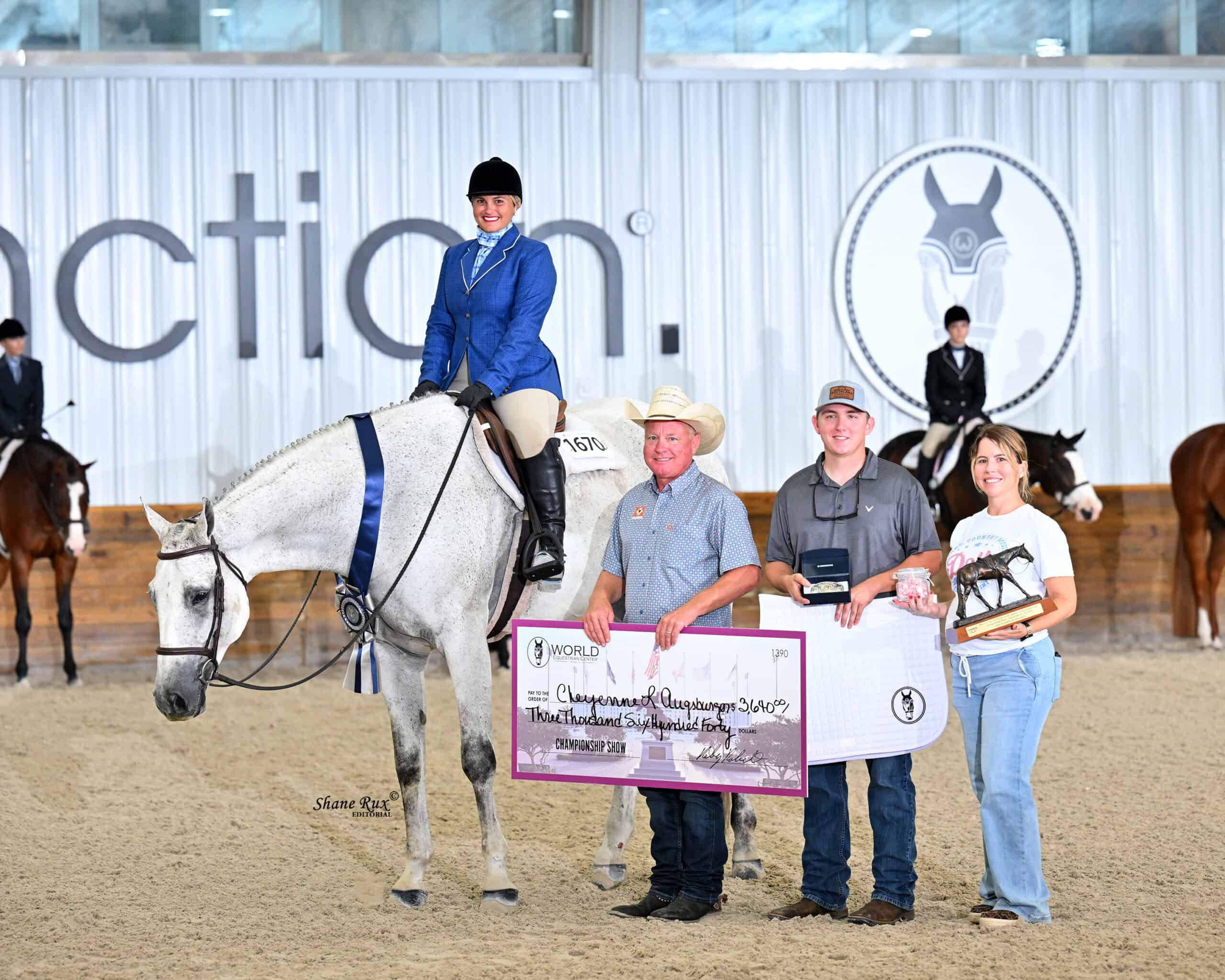 A woman on a white horse poses with three people holding a large check, plaques, and a trophy in an indoor arena. Other riders and horses are seen in the background.