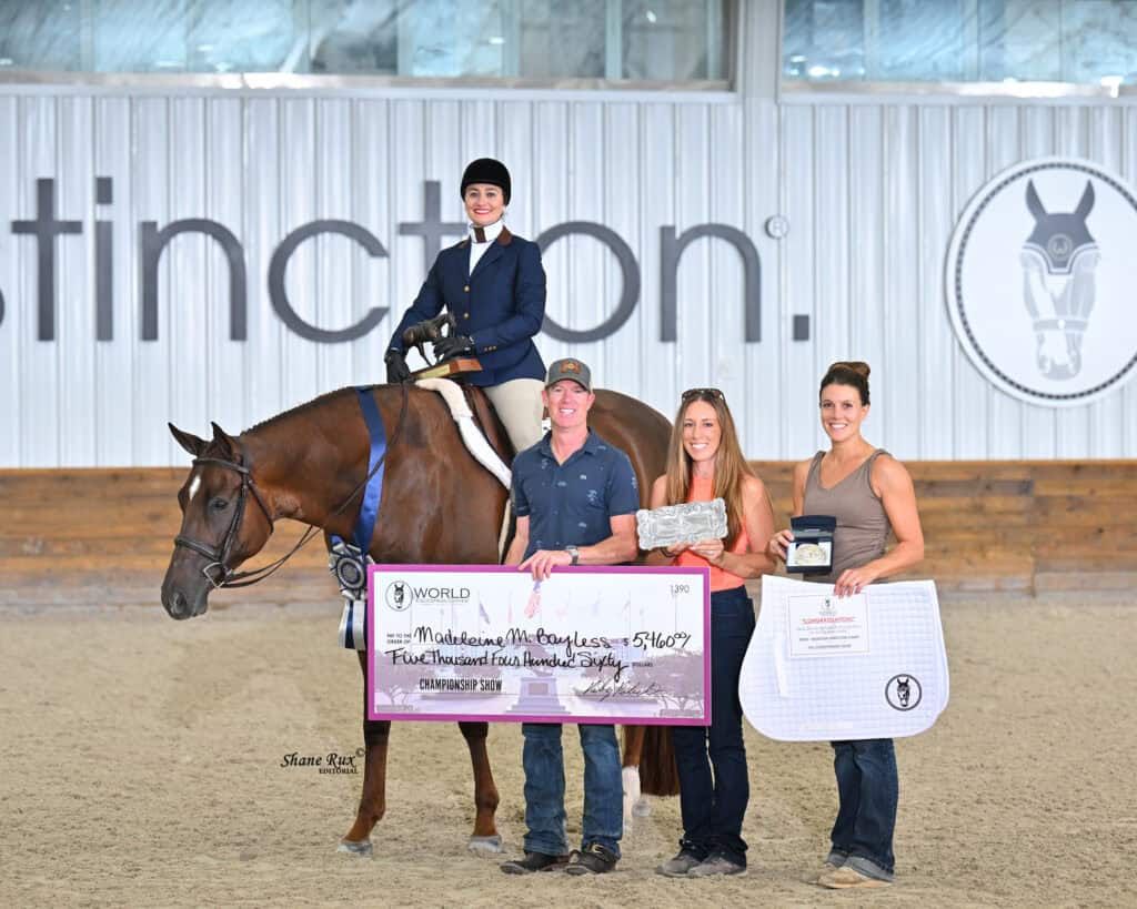Four people and a horse pose indoors; one person is on the horse in riding attire, while the others hold a large check, trophy, and equestrian gear.