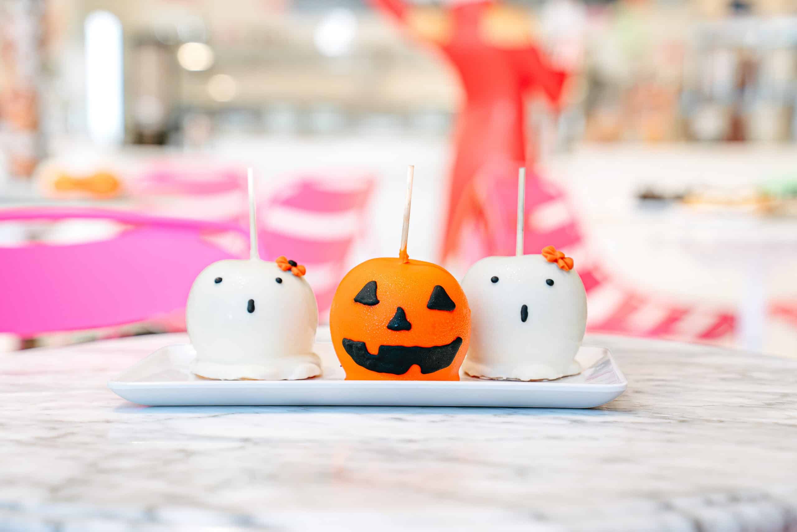 Three candy apples decorated as two white ghosts and an orange jack-o’-lantern sit on a white rectangular plate on a marble table.