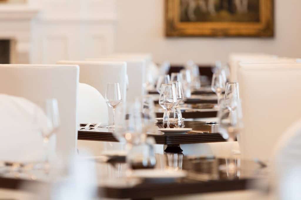 A close-up view of several dining tables set with glassware, plates, and cutlery in a bright, elegant restaurant with white chairs.