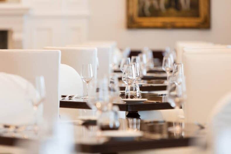 A close-up view of several dining tables set with glassware, plates, and cutlery in a bright, elegant restaurant with white chairs.