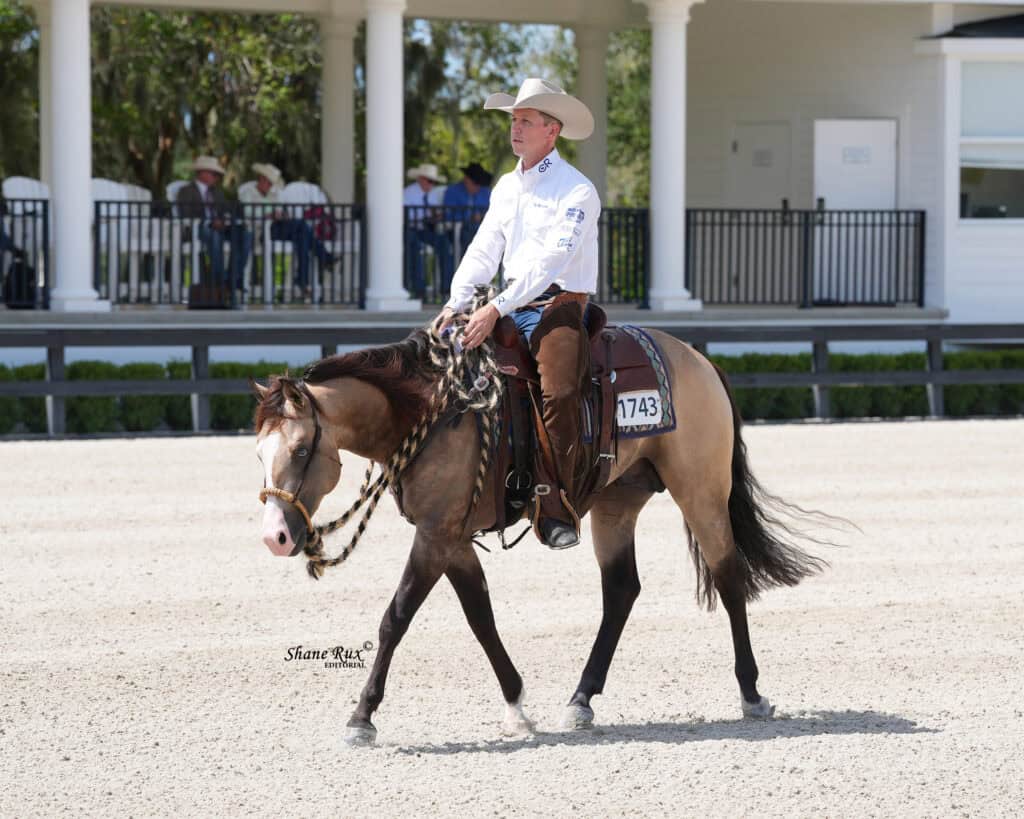 A man in a cowboy hat rides a horse at an outdoor equestrian event, with a white building and spectators in the background.