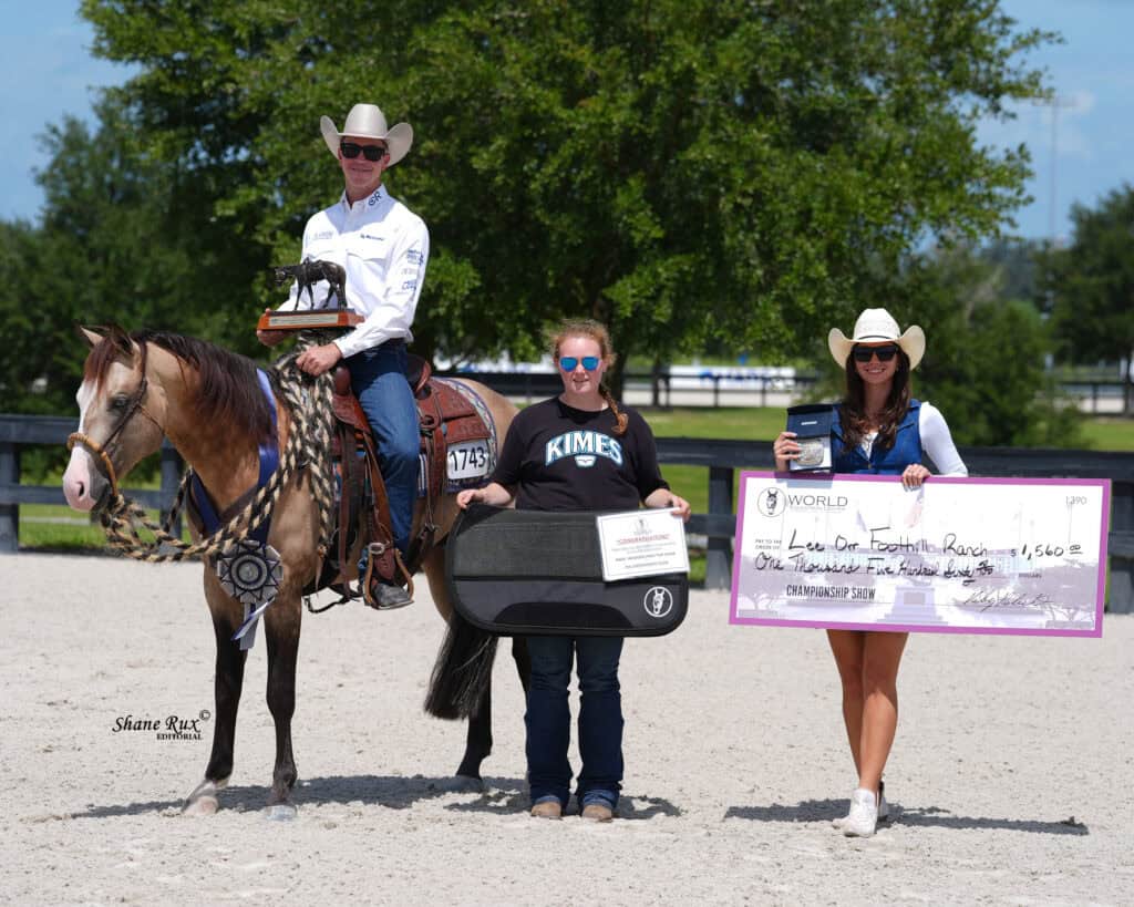 Three people and a horse pose outdoors; one person is on horseback, another holds an award case, and the third holds a large check for $1,560.
