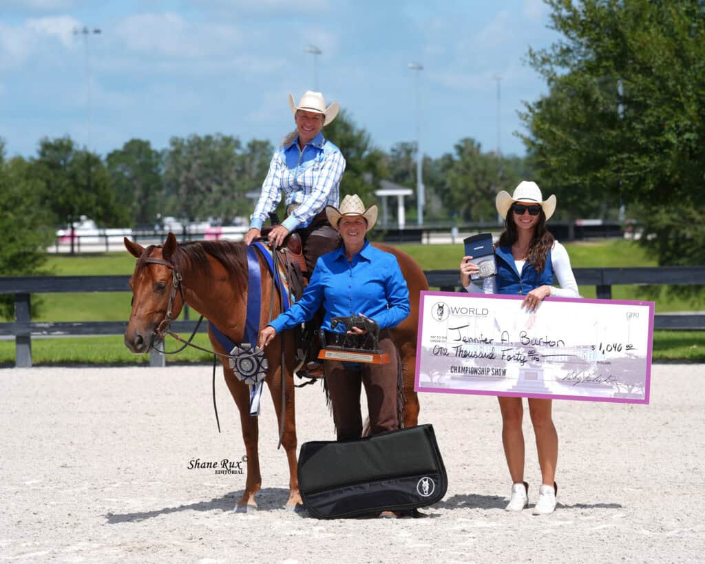 Three people in western attire pose outdoors with a horse, a trophy, a large check, and an award plaque.