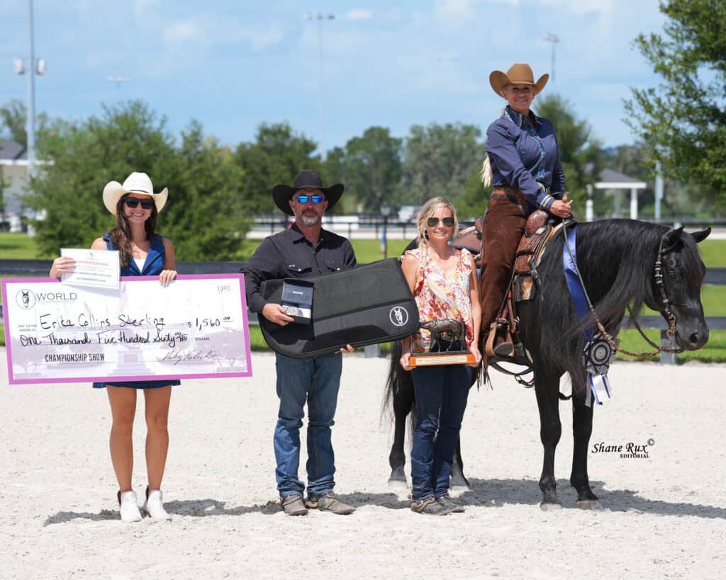 Four people pose outdoors, one on a dark horse. They hold a large check, a trophy, and a case. All are smiling and dressed in Western-style clothing and hats.
