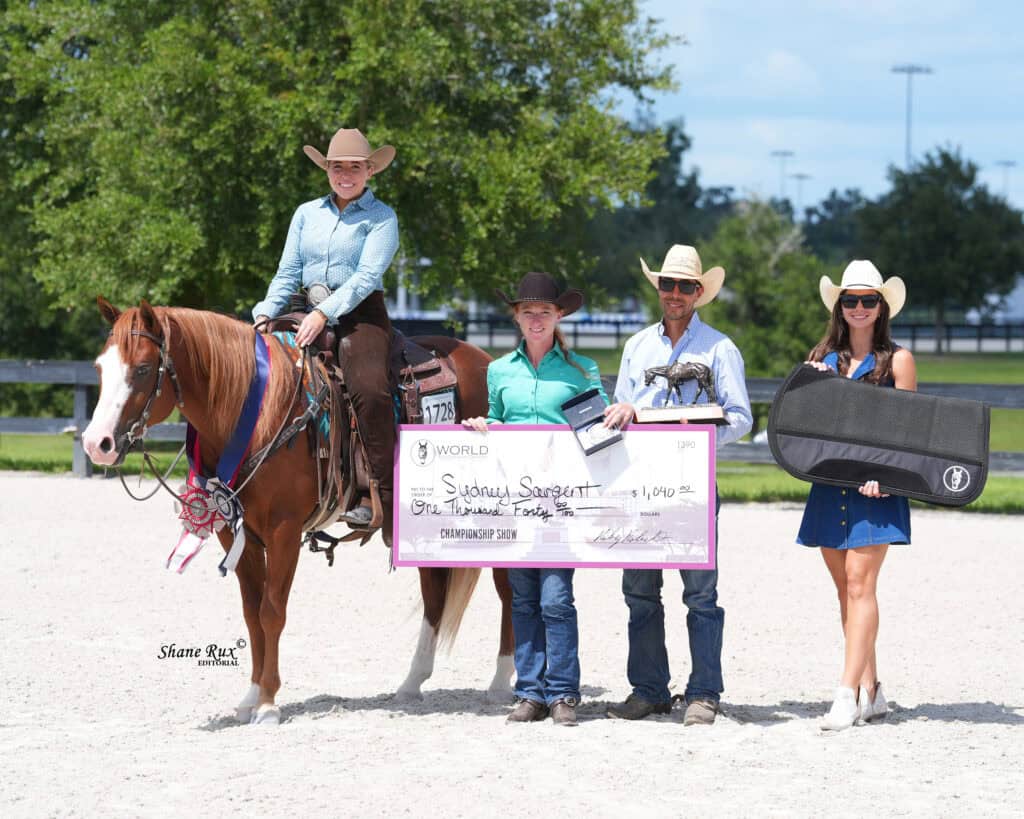 Four people in cowboy hats pose outdoors with a horse, holding a large check and a guitar case. One person is seated on the horse while the others stand on the ground.