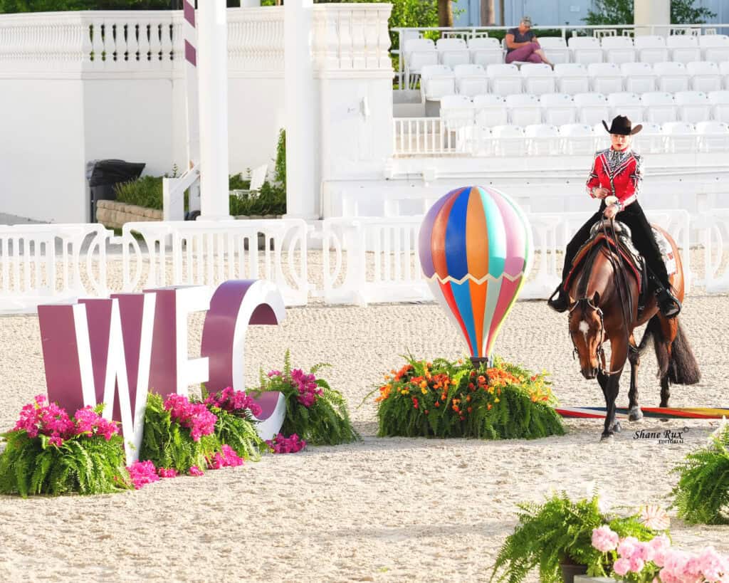 A rider in a red jacket and black hat rides a horse past a “WEC” sign and a colorful hot air balloon decoration in an outdoor arena.