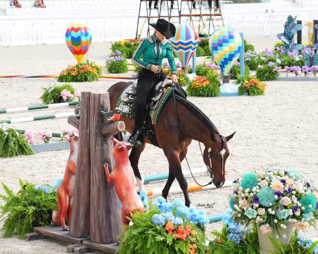 A rider in a green shirt and black hat guides a horse around colorful obstacles and flower arrangements in an equestrian arena.