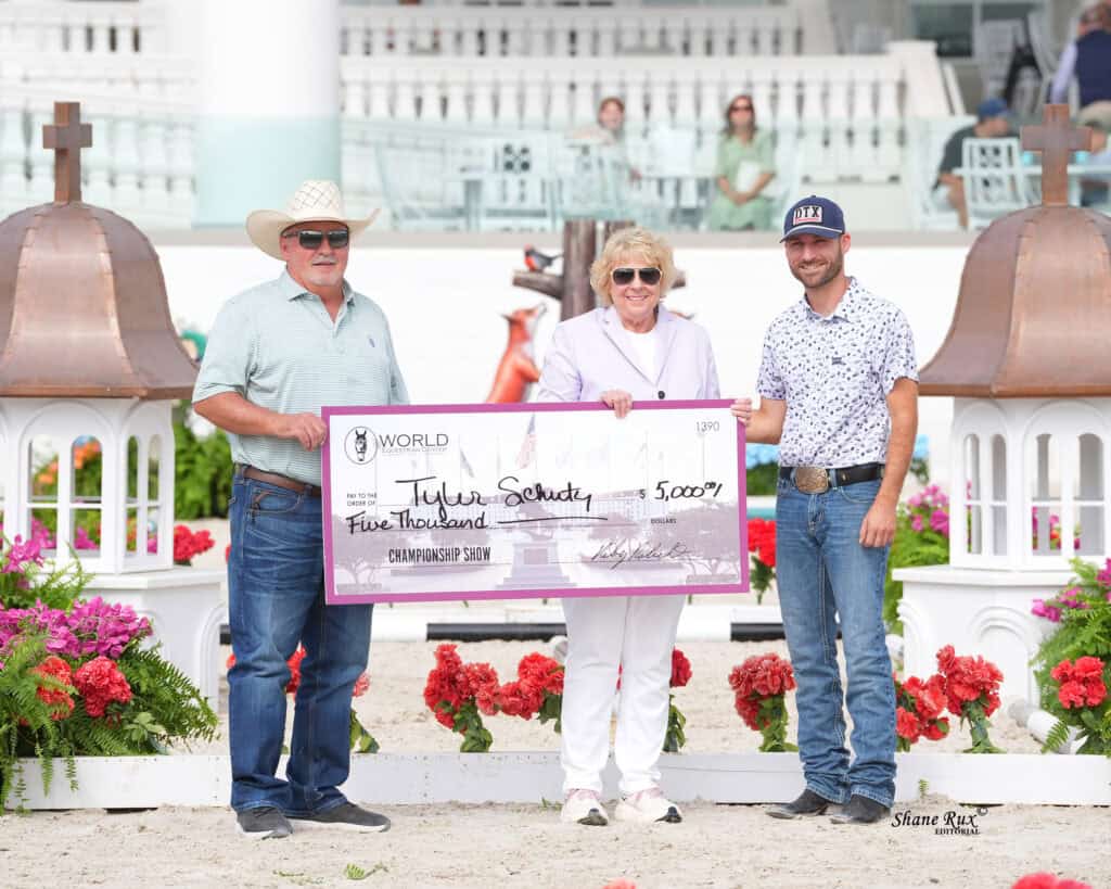 Three people stand outdoors holding an oversized check for $5,000. Flowers and small white structures are in the background.