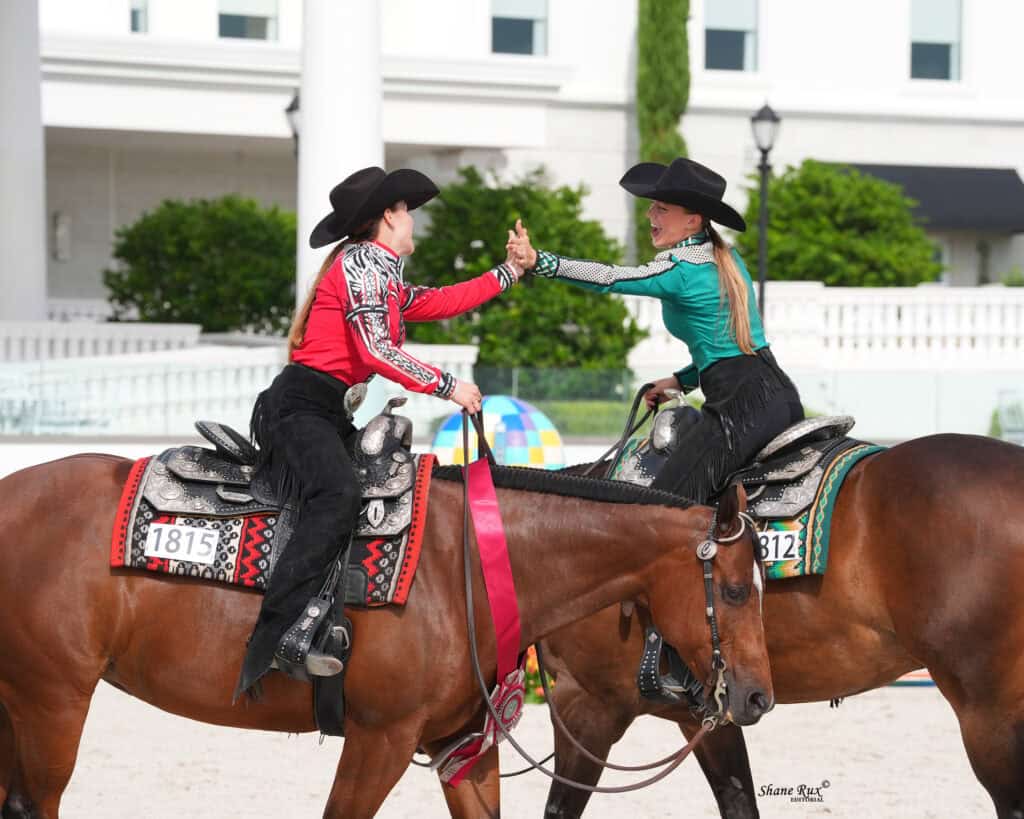 Two women in western attire riding horses give each other a high five during a competition. One horse has a red ribbon. Background includes greenery and a white fence.