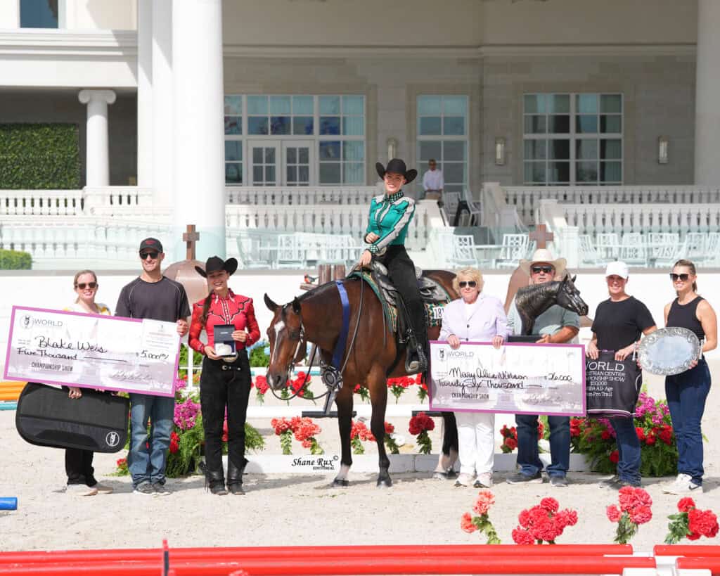 A group of people, including a rider on a horse, pose outdoors with large award checks, flowers, and trophies in front of a white building.