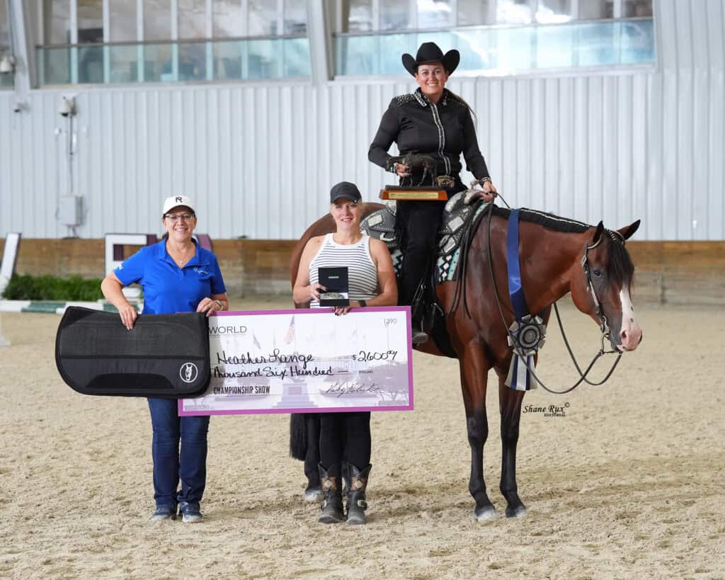 Three women pose indoors at a horse event; one is on horseback holding a trophy, while the other two stand beside her holding a large check and an award.