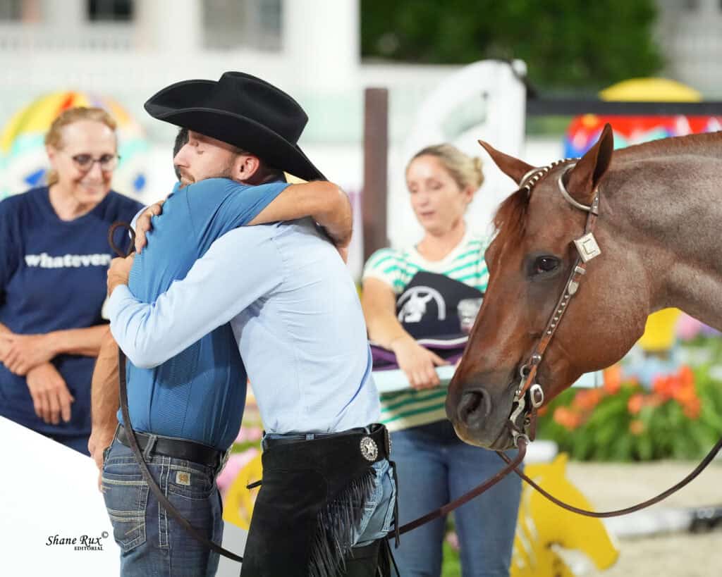 Two men hug while a brown horse stands nearby; two women are in the background, one wearing a striped shirt and the other a blue shirt.