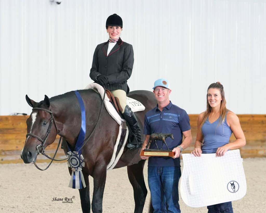 A rider on a horse with a blue ribbon poses next to two people holding a trophy and a white saddle pad in an indoor arena.