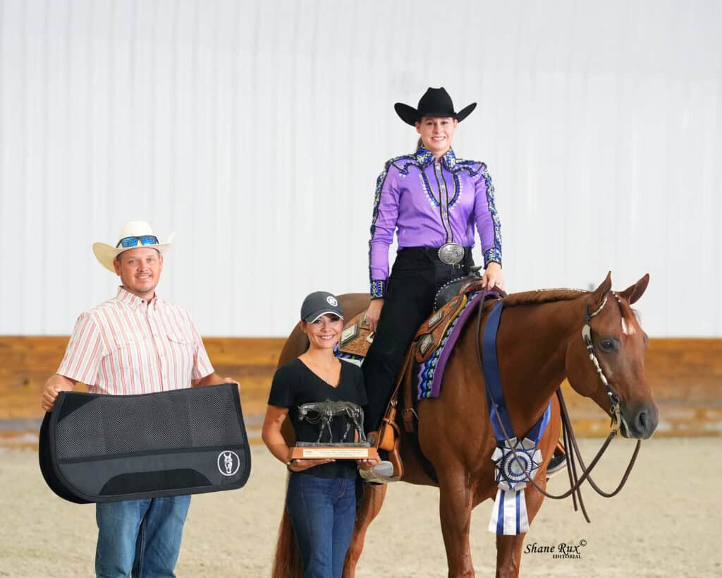 Three people pose indoors with a horse; the rider wears a purple shirt and holds a ribbon, while the others hold a case and a trophy.