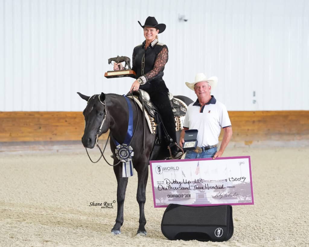 A woman in western attire sits on a horse holding a trophy, next to a man holding a large $13,000 check, with a championship ribbon and award case visible.