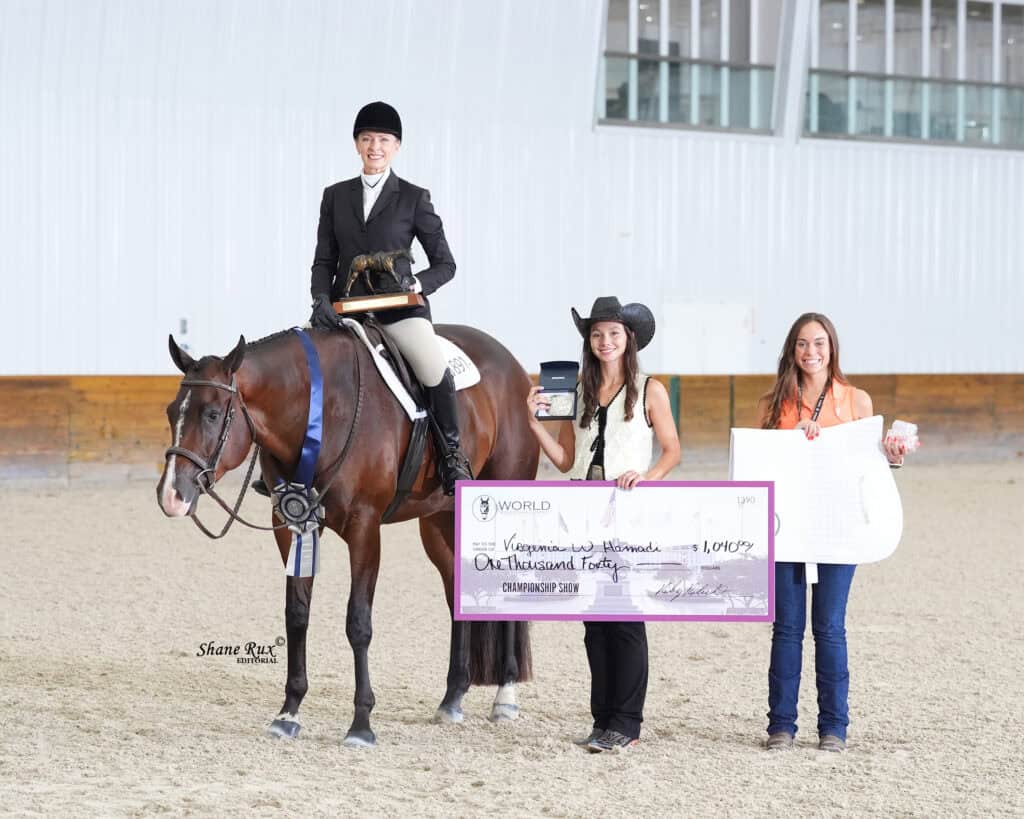 Three women pose indoors, one on horseback in riding attire, two standing and smiling while holding a large championship check and ribbon; arena setting with wood and glass walls.