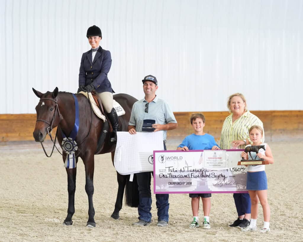A woman on horseback poses with three adults and two children holding a large check and ribbon in an indoor arena.