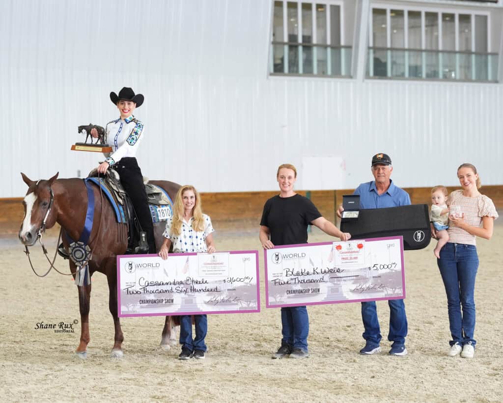 A woman on horseback and four people stand indoors holding two oversized checks and a belt buckle, with a baby held by one adult.