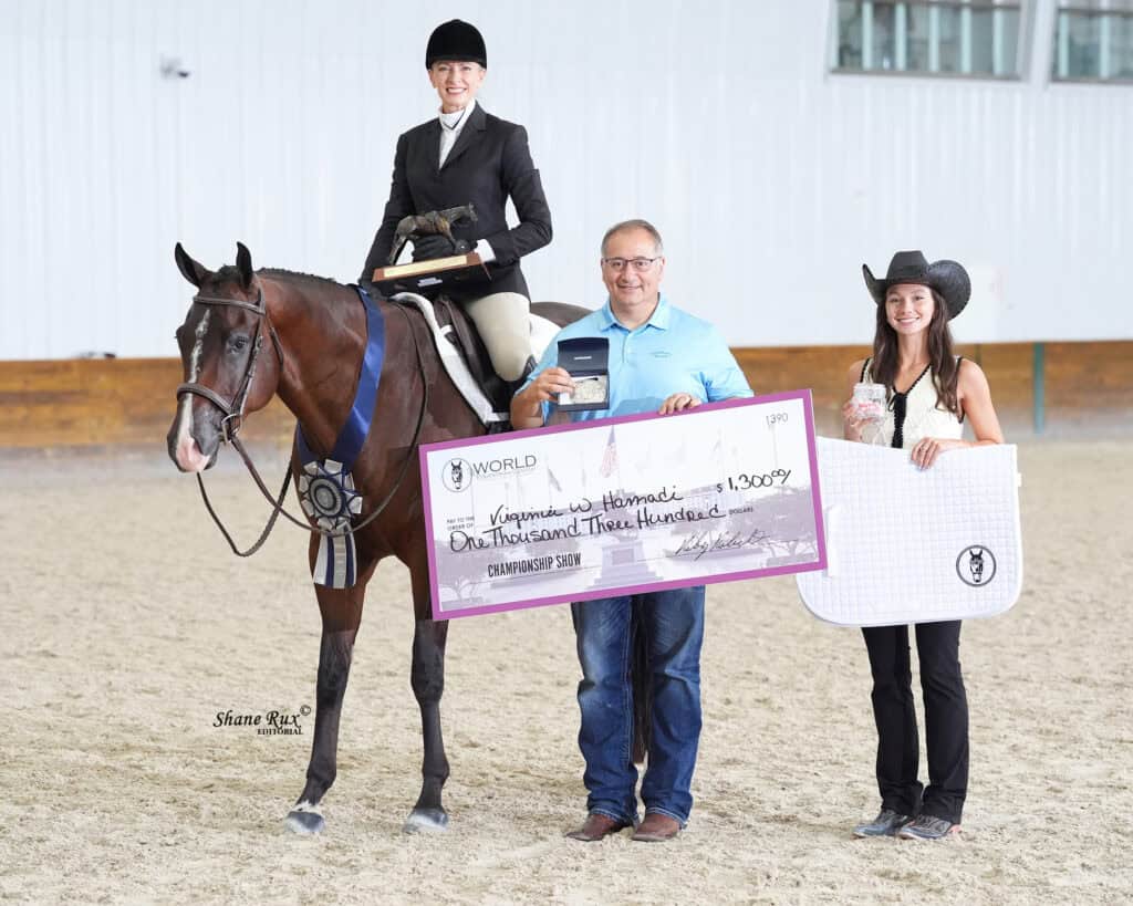 A woman on horseback, a man holding an oversized check for $1,300, and another woman in western attire stand together in an indoor arena.