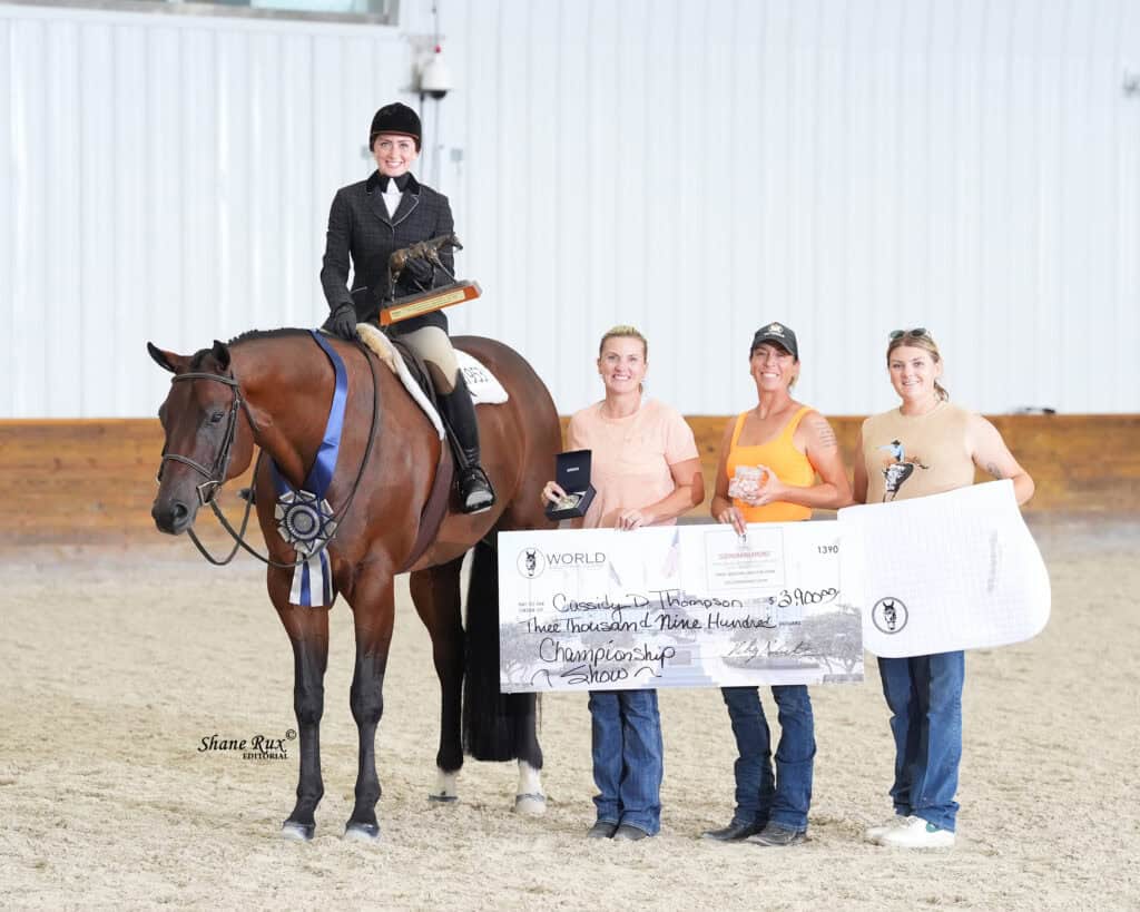 A rider on a horse with ribbons poses indoors, while three people beside them hold a large championship check, medals, and prizes.