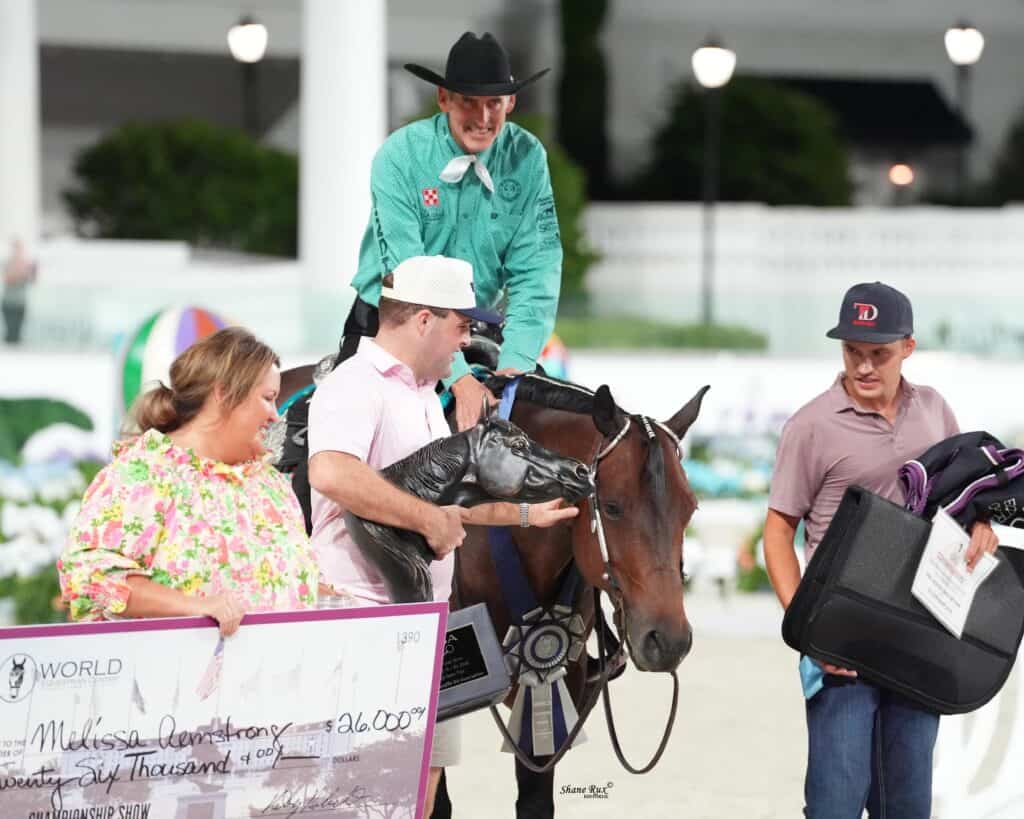 A man on horseback smiles while others stand nearby holding a large check, a trophy, and horse equipment at an indoor equestrian event.