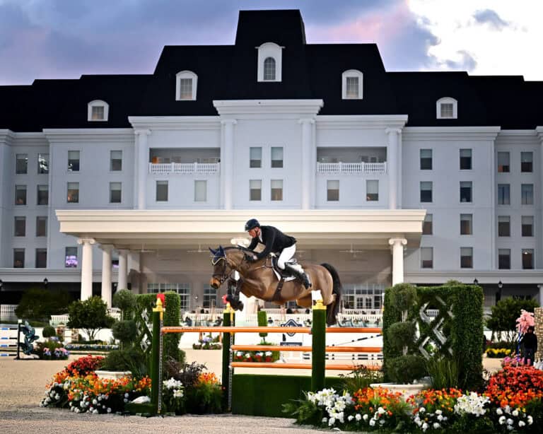 Equestrian rider in black attire jumps a horse over a green and orange obstacle in front of a large white building, surrounded by flowers and greenery.