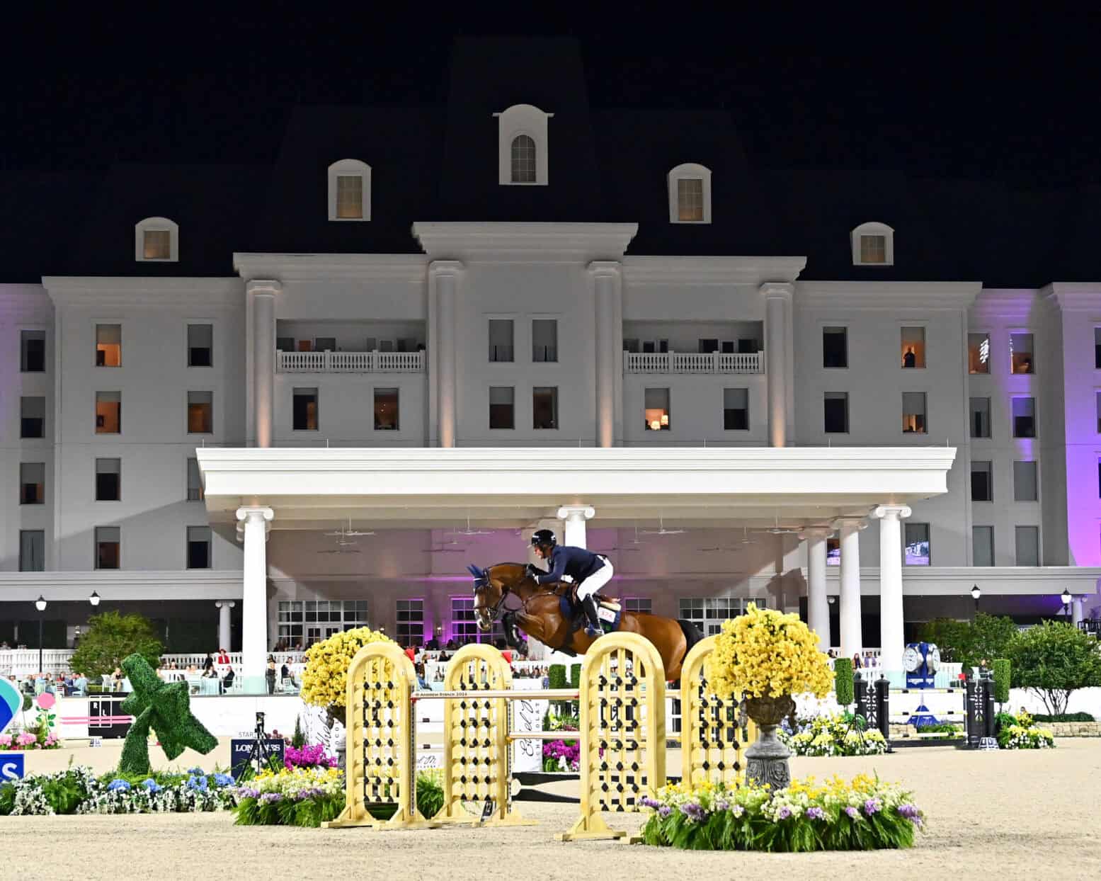A horse and rider jump over an obstacle during an equestrian event at night in front of a large, lit hotel building.
