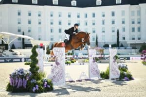 A rider in a black jacket and helmet jumps a brown horse over a decorative obstacle at an outdoor equestrian event, with a large white building in the background.