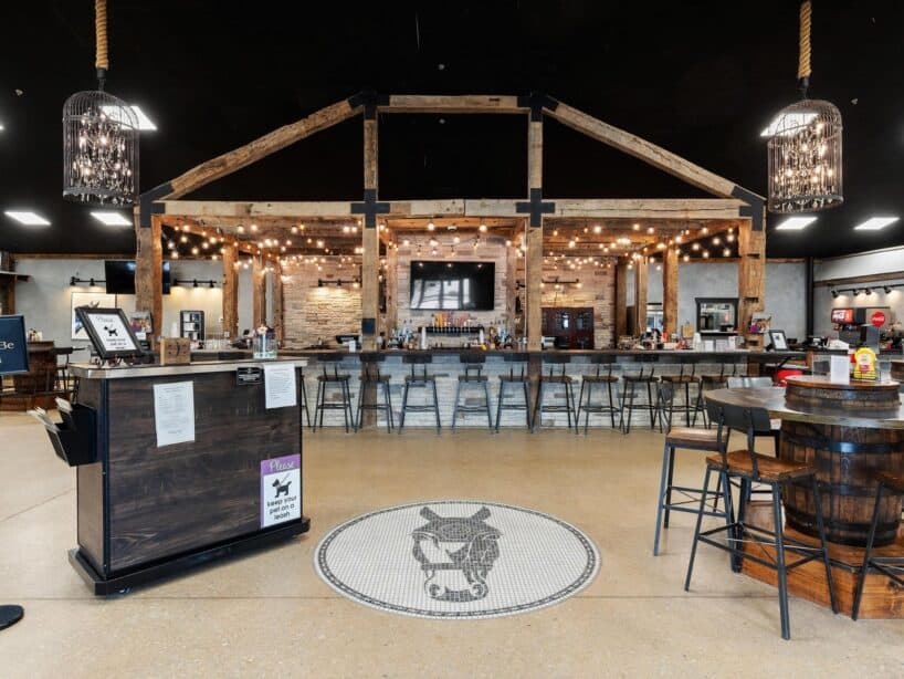 A rustic bar with wooden beams, string lights, a large TV above the counter, and a donkey mosaic on the floor. Tables and chairs are arranged throughout the spacious room.
