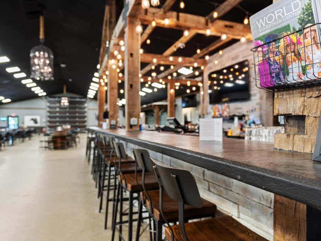 Long wooden bar with stools in a spacious, modern restaurant featuring exposed beams, string lights, and a magazine rack on the countertop.