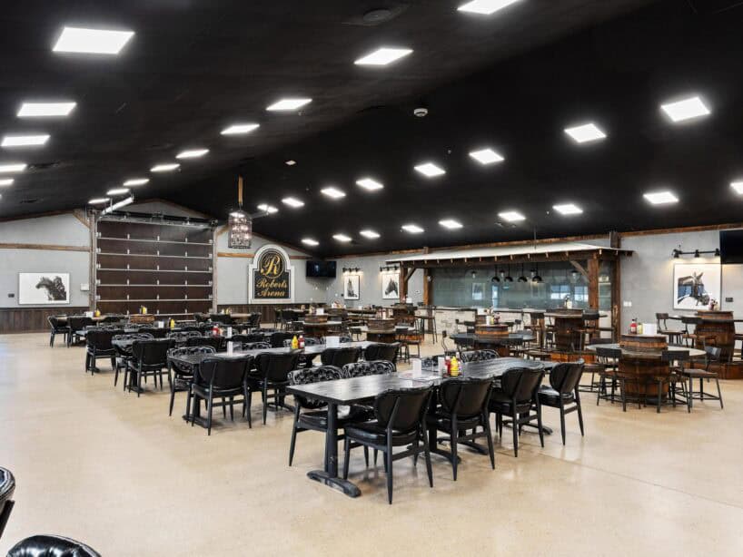Spacious restaurant dining area with black chairs and tables, light-colored floor, high ceiling with bright lights, and western-themed decor on the walls.