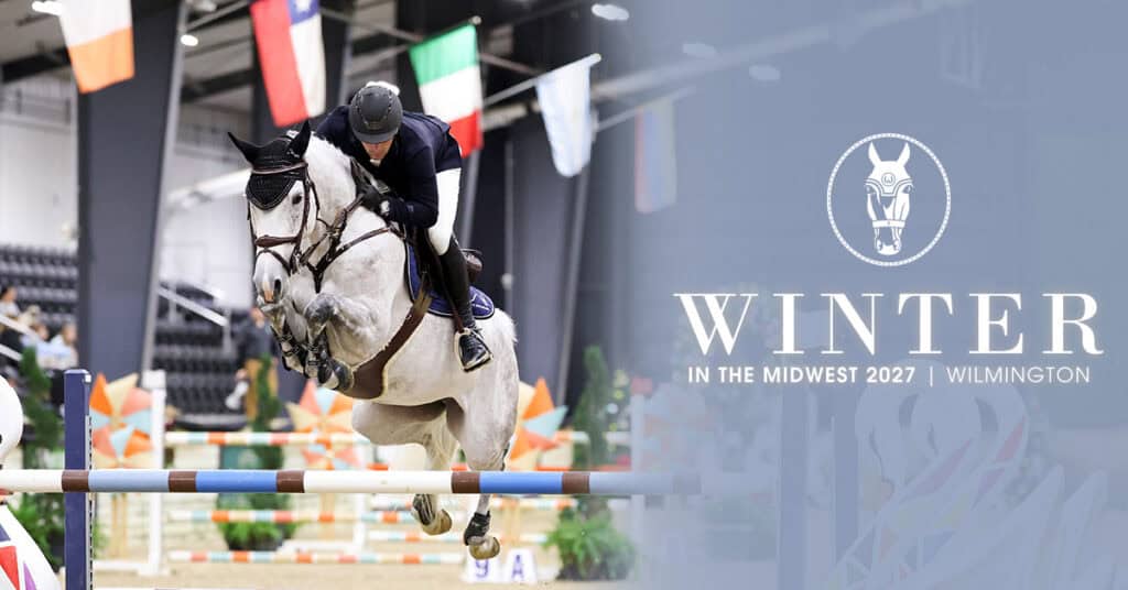 A rider and horse clear a jump during an indoor equestrian event, with event branding that reads “Winter in the Midwest 2027 Wilmington” on the right.