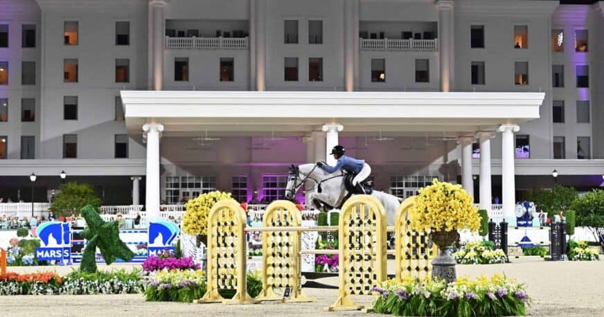 A horse and rider jumping over an obstacle during an equestrian competition in front of a large white building.