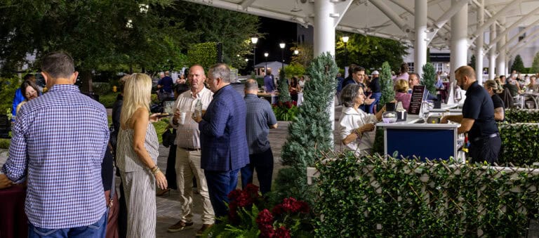 People gather under a covered outdoor area, socializing near a bar and greenery at night, with bartenders serving drinks and trees in the background.