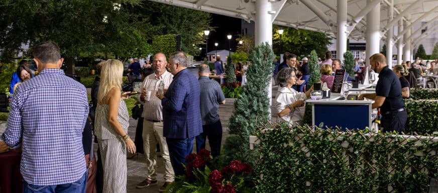 People gather under a covered outdoor area, socializing near a bar and greenery at night, with bartenders serving drinks and trees in the background.