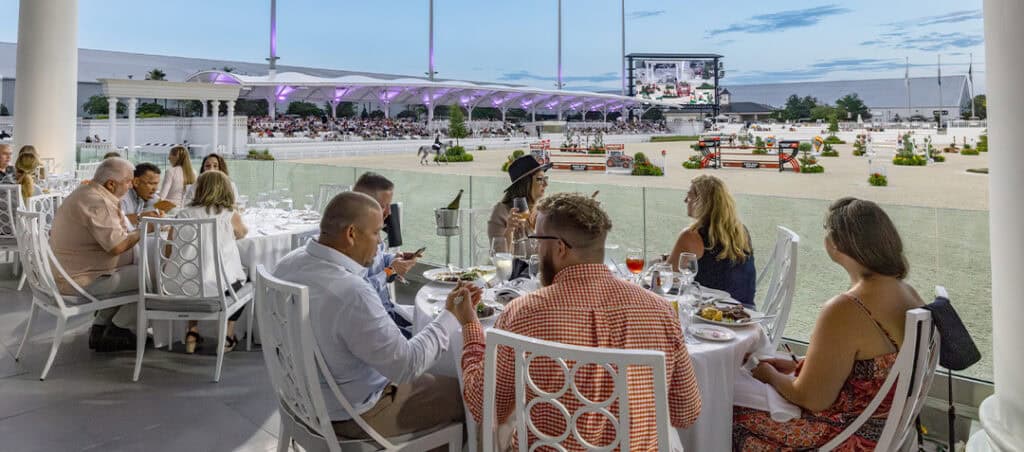 People dine at outdoor tables overlooking an equestrian arena, where a horse jumping event is taking place under stadium lights at dusk.