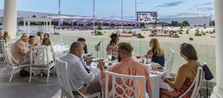 People dine at outdoor tables overlooking an equestrian arena, where a horse jumping event is taking place under stadium lights at dusk.