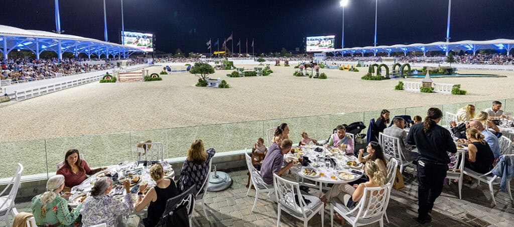People dine at outdoor tables overlooking a well-lit equestrian arena at night, with spectators seated in stands and horse jump obstacles visible in the arena.