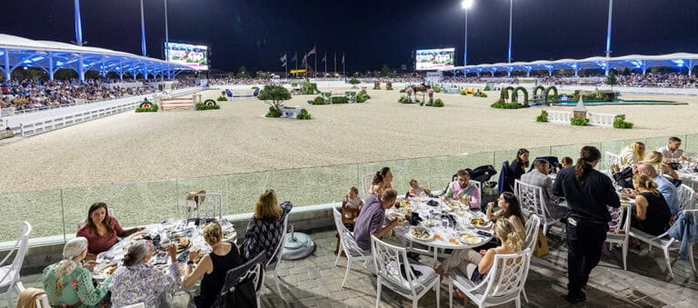 People dine at outdoor tables overlooking a well-lit equestrian arena at night, with spectators seated in stands and horse jump obstacles visible in the arena.