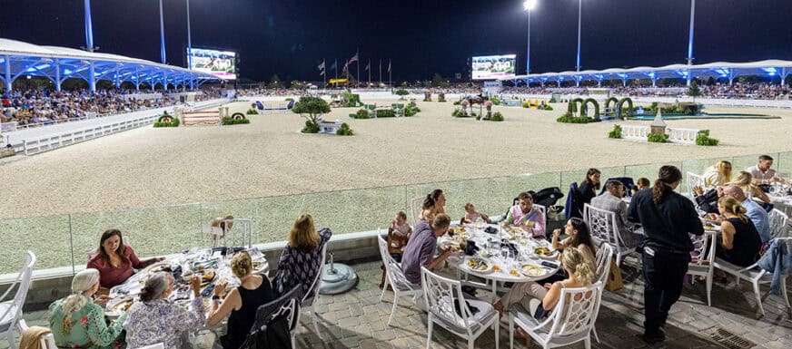 People dine at outdoor tables overlooking a well-lit equestrian arena at night, with spectators seated in stands and horse jump obstacles visible in the arena.