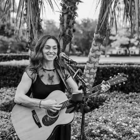 A woman is playing an acoustic guitar and singing into a microphone outdoors, surrounded by plants and palm trees.