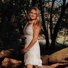 A woman in a white dress stands outdoors in a wooded area, with sunlight filtering through the trees.