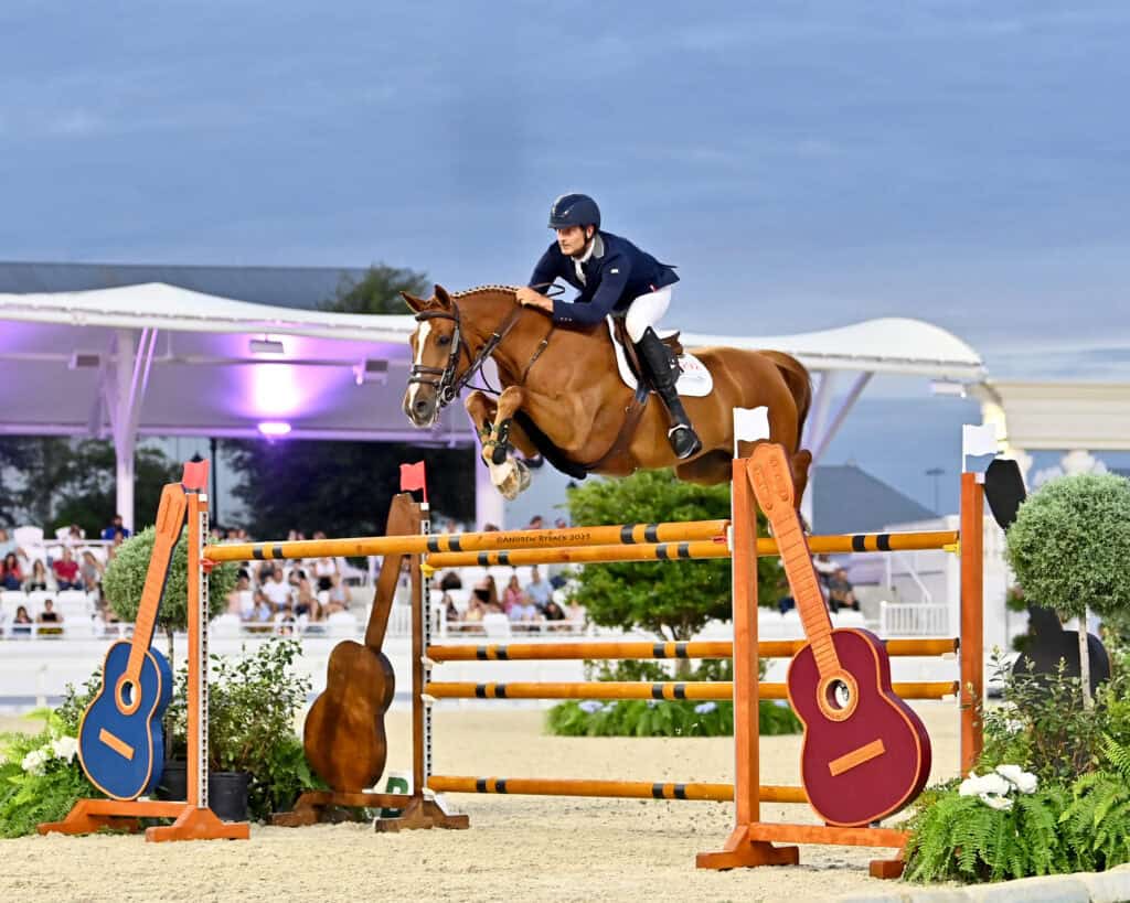 Equestrian rider on a brown horse jumps over a high obstacle decorated with large guitar shapes during a competition.