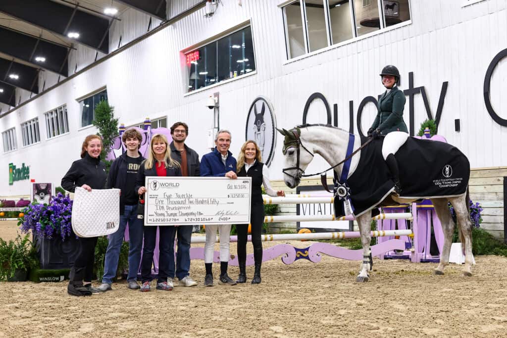 A rider on a grey horse stands next to five people holding a large ceremonial check for $1,250.00 at an indoor equestrian arena.