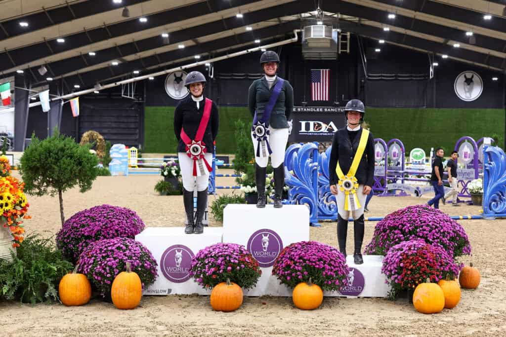 Three equestrians in riding attire standing on a winners’ podium decorated with purple chrysanthemums and pumpkins.